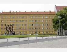 Gelbes, mehrstöckiges Wohngebäude mit einem roten Ziegeldach, davor eine Rasenfläche mit einer großen Metallskulptur in Form eines Rades. Im Vordergrund ist ein gepflasterter Platz und eine Straße mit parkenden Autos zu sehen.