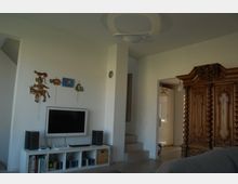 Wohnzimmer mit modernen und klassischen Elementen, darunter ein Flachbildfernseher auf einem weißen Regal, eine hölzerne Wandverzierung und ein großer, reich verzierter antiker Kleiderschrank. Im Hintergrund führt eine kleine Treppe zu einem angrenzenden Raum.