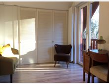 Ein heller, schlicht eingerichteter Raum mit Holzboden, einem Sessel in der Mitte, einem Schreibtisch mit Stuhl rechts und einer Sitzbank mit gelbem Kissen links. Große Glastüren mit Blick auf einen Balkon und Bäume lassen Tageslicht herein, daneben sind weiße Lamellentüren zu sehen.