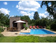 Garten mit einem rechteckigen Pool, umgeben von gepflasterten Flächen und fünf roten Liegestühlen. Links befindet sich eine kleine Backsteingebäude mit Rundbogenfenster und Holztischen und -stühlen unter einem Sonnenschirm, umgeben von üppigem Grün und Bäumen.