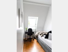 Kleines Schlafzimmer mit Dachschräge, einem Holzboden, einem schmalen Bett mit grauem Stoffrahmen, und einem kleinen Schreibtisch mit schwarzem Stuhl unter einem Dachfenster. An der Wand hängen zwei Bilder, und eine Pflanze steht auf dem Schreibtisch.