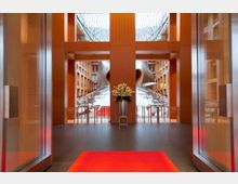 Lobby eines modernen Gebäudes mit warmen Holztönen, einer roten Teppichläufer in der Mitte und einem auffälligen, skulpturalen Architekturdetail im Hintergrund. Große Glasflächen und ein gewölbtes Glasdach lassen viel Tageslicht herein.