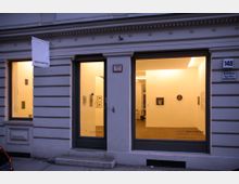 Fassade einer Kunstgalerie mit zwei großen Schaufenstern, durch die ein beleuchteter Ausstellungsraum mit Bildern an den Wänden sichtbar ist. Über dem Eingang hängt ein Schild mit der Aufschrift „Galerie Martin Mertens“, daneben befindet sich die Hausnummer 148.