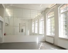 Heller, leerer Innenraum im Loft- oder Atelierstil mit hohen Decken, grauem Boden und einer großen Glas-Trennwand mit Metallrahmen. An der rechten Seite stehen mehrere hohe Sprossenfenster mit Rundbögen, darunter Heizkörper; an der Decke sind lange Leuchtstofflampen montiert.