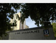 U-Bahn-Eingang "Hansaplatz" mit Schriftzug und U9-Symbol auf gefliester Wand, im Hintergrund ein großes Wohnhochhaus. Der obere Bildrand ist von Laubbaumzweigen eingerahmt.