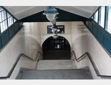 Treppenabgang in einer U-Bahn-Station mit weißen Fliesenwänden, metallenen Handläufen und einem gewölbten Durchgang. Über dem Ausgang hängt ein Schild mit der Aufschrift "Welcome to Station Berlin".