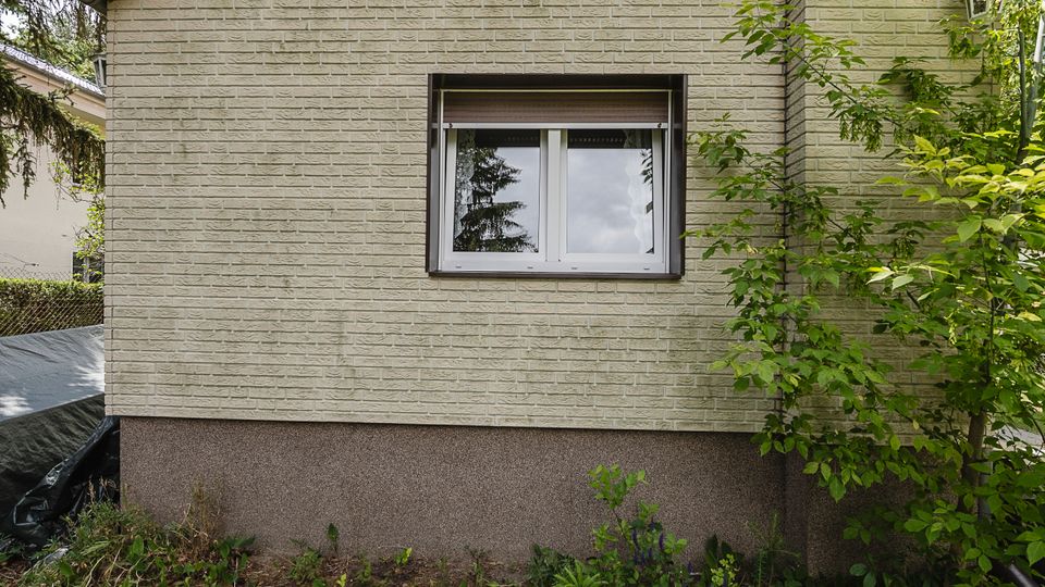 Außenwand eines Wohnhauses mit hellen Klinkersteinen und einem zentralen Doppelfenster mit Rollladen. Unten ist ein dunkler Sockelputz zu sehen; davor wachsen Sträucher und Gartenpflanzen.