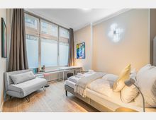 Helles, modern eingerichtetes Schlafzimmer mit großem Bett, kleiner Sitzbank und Schreibtisch vor einer breiten Fensterfront mit dunklen Vorhängen. Auf hellem Holzfußboden stehen ein kleiner Beistelltisch und eine Stehlampe; an der Wand hängt ein Poster und eine geometrische Wandleuchte.