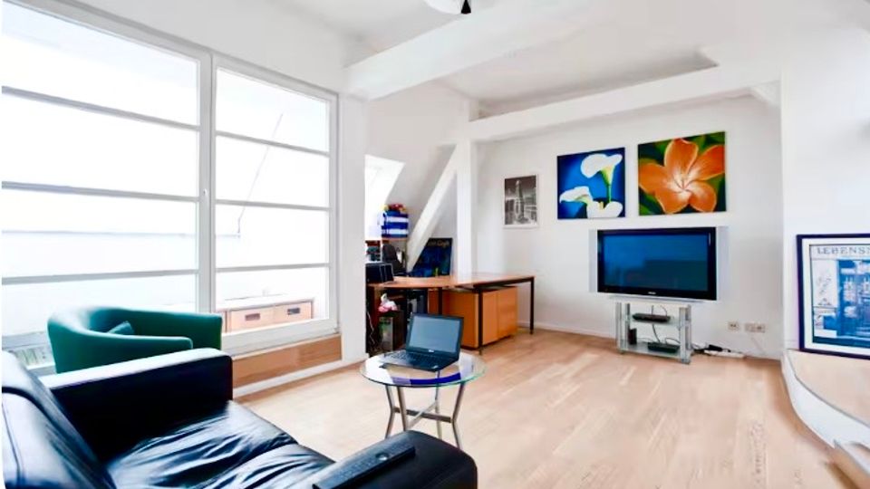 Heller Wohnbereich in einer Dachgeschosswohnung mit großen Fensterfronten, hellem Holzfußboden und weißen, leicht schrägen Deckenbalken. Links stehen Sofa und Sessel, mittig ein kleiner Glastisch, rechts ein Fernseher mit Lowboard und Wandbildern darüber sowie ein Arbeitsbereich mit Tisch.