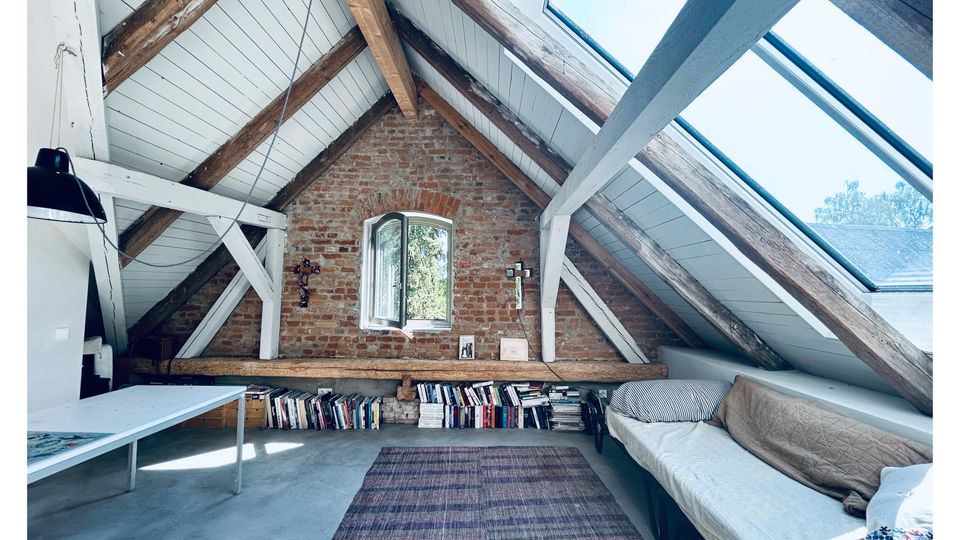 Dachgeschoss-Schlafzimmer mit schrägem, weiß verkleidetem Dach, sichtbaren Holzbalken und großen Dachfenstern, die viel Tageslicht hereinlassen. An einer Backsteinwand mit kleinem Fenster stehen hohe Bücherstapel; daneben sind Bett und Teppich in einem schmalen Raum angeordnet.