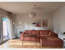 Wohnzimmer mit einem Ecksofa in Terrakottafarbe, einem kleinen Holztisch und einem hölzernen Fischgrätenparkettboden. An der Wand hängen zwei gerahmte Poster, daneben steht eine Stehlampe mit mehreren kugelförmigen Lampenschirmen. Links führen bodentiefe Fenster mit weißen Vorhängen auf eine Terrasse.