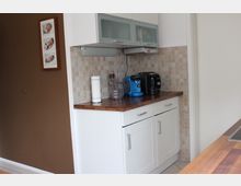 Weiße Küche mit Holzarbeitsplatte, auf der zwei Kaffeemaschinen und ein Kapselständer stehen. Die Rückwand besteht aus beigefarbenen Fliesen, und an der angrenzenden braunen Wand hängt ein Bilderrahmen mit Fotos.