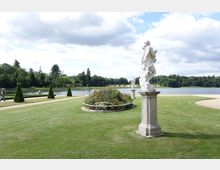 Gepflegter Park mit Rasenflächen, Blumenbeeten und symmetrisch angeordneten Statuen auf Steinsockeln. Im Hintergrund ist ein ruhiger See mit umgebenden Bäumen unter einem bewölkten Himmel zu sehen.