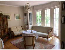 Wohnzimmer mit klassischem Design, ausgestattet mit einem Holzboden, gepolsterten Sofas, einem weißen Tisch mit Kerzenhaltern, einem orientalischen Teppich und einem holzgeschnitzten Schrank. Große Fenster mit Sprossen lassen viel Tageslicht herein.