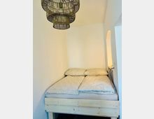 Kleines Schlafzimmer mit einem schmalen Doppelbett in einer Nische, weißen Wänden und hellem Holz-Bettgestell. An der Decke hängt eine geflochtene Rattanlampe; seitlich befindet sich ein arched Wanddurchbruch mit indirekter Beleuchtung.