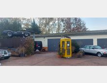 Hof mit einfacher Garage, davor mehrere Fahrzeuge einschließlich eines alten Sportwagens auf einem Container. Rechts steht eine gelbe Telefonzelle, umgeben von Pflanzen und Bäumen im Hintergrund.