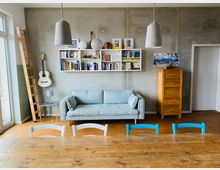 Wohnzimmer mit hellgrauer Couch, hölzernem Esstisch und Stühlen in Blau und Weiß. An der Wand befinden sich ein Bücherregal, eine Gitarre, Bilderrahmen und eine Leiter; daneben steht eine Holzkommode.