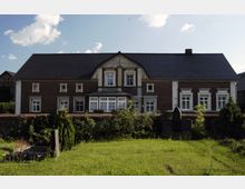 Backsteinhaus mit dunklem Ziegeldach und weißen Fensterrahmen, in der Mitte ein verglaster Vorbau. Im Vordergrund eine Rasenfläche mit Grabsteinen und einer Hecke, die das Haus vom Garten abgrenzt.