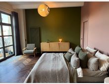 Modern bedroom with a large bed covered in neutral bedding and multiple pillows, set against a muted pink wall and a dark green accent wall. A wooden sideboard, tall radiator, gray armchair, and pendant light sit on herringbone wood flooring beside a large glass door to a small balcony.