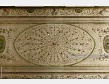 Deckenansicht eines historischen Innenraums mit kunstvollen Stuckverzierungen, floralen Ornamenten und einem symmetrischen Muster, das von einem ovalen Rahmen eingerahmt ist. Die Farbgebung ist überwiegend in gedeckten Tönen mit Grün- und Beigetönen gehalten.