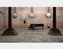 Industrieller Innenraum mit einer freigelegten Ziegelwand, zwei Metallpfeilern und einem Flügel, der mit einer schwarzen Stoffhülle abgedeckt ist. Über dem Klavier hängen große, runde Lampen an höhenverstellbaren Halterungen von der Decke.