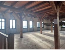 Innenraum eines historischen Gebäudes mit Holzbalken, Holzdach und freistehenden Holzsäulen. Der Raum hat große, bogenförmige Fenster und einen Holzboden, durch den Tageslicht einfällt.