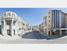 Straßenszene mit symmetrisch angeordneten, historischen Gebäuden im Stil der klassischen Architektur, darunter Rundbalkone mit Säulen und dekorativen Fassaden. Die Straße ist leer, und die Umgebung wirkt wie ein Filmset oder eine künstliche Kulisse unter klarem, blauem Himmel.