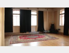 Ein schlichter Raum mit Holzboden, großen Fenstern mit schwarzen Vorhängen und einem roten, gemusterten Teppich in der Mitte. In einer Ecke sind gestapelte Schlagzeugteile und Zubehör zu sehen.