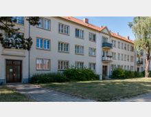 Wohngebäude mit drei Stockwerken und ziegelrotem Satteldach, großen rechteckigen Fenstern und Balkonen. Das Gebäude ist von Grünflächen, Büschen und Bäumen umgeben, ein gepflasterter Gehweg führt zum Eingang.