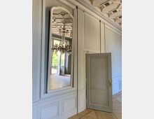 Innenraum eines historischen Gebäudes mit einer hohen weißen Wand, die durch vertikale Zierleisten gegliedert ist. Ein großer Wandspiegel spiegelt einen Kronleuchter, Kassettendecken und ein helles Zimmer mit Blick nach draußen, daneben eine geöffnete Holzinnentür.