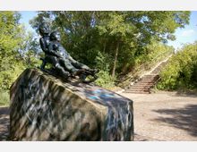 Bronzestatue von zwei Kindern auf einem Schlitten, die auf einem großen, bemalten Sockel steht, umgeben von Bäumen und einer Treppenanlage im Hintergrund. Die Szene befindet sich in einem parkähnlichen Außenbereich mit gepflastertem Boden.