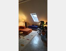 Wohnraum unter einer Dachschräge mit einem Dachfenster, das Tageslicht hereinlässt. Der Raum enthält ein oranges Sofa, einen bunten Teppich, eine Stehlampe, einen Schreibtischstuhl, eine Zimmerpflanze auf einem Beistelltisch und ein blaues Sideboard.