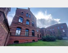 Backsteinbau im historischen Stil mit großen Rundbogenfenstern, dekorativen Backsteinelementen und Erkern; im Vordergrund eine Rasenfläche, umgeben von wenigen Büschen, unter einem bewölkten Himmel.