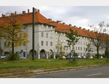 Wohnblock einer historischen Siedlung mit hellen Fassaden und roten Ziegeldächern, unterbrochen von Arkaden mit Graffiti. Im Vordergrund befinden sich eine Straßenbahntrasse, geparkte Autos, Bäume und eine Grünfläche.