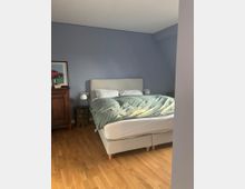 Schlafzimmer mit einem grauen Boxspringbett auf hellem Holzboden. Eine blaue Wand im Hintergrund, links ein Holzschrank mit einem gerahmten Bild darauf und zwei kleine Nachttische mit Lampen daneben.
