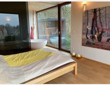 Schlafzimmer mit Holzboden und einem Holzbett, bedeckt mit einer gestreiften Decke in Gelb- und Beigetönen. Ein großes Fenster mit Blick auf eine Terrasse und Bäume, daneben eine freistehende Badewanne; an der Wand hängt ein Bild eines Waldes.