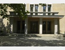 Eingangsbereich eines Gebäudes mit hellen Steinwänden, rechteckigen Fenstern und einer Tür mit Gittermuster. Der Eingang wird von einem Vordach mit massiven Steinsäulen gestützt, umgeben von gepflastertem Boden und teilweise durch Baumkronen beschattet.