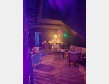 Gemütliche Lounge mit gedämpfter Beleuchtung in violetten und warmen Gelbtönen. Der Raum verfügt über Polstersessel, Sofas, Holzboden sowie einen Bogen-Durchgang und wird von einer Stehlampe beleuchtet.