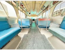 Innenraum eines klassischen Busses mit zwei Reihen aus fest installierten, überwiegend blauen Sitzen auf beiden Seiten und einem Mittelgang. Die Wände und Fenster sind leicht gewölbt, und die Decke zeigt eine beige Verkleidung mit Metallstangen zur Stabilisierung.