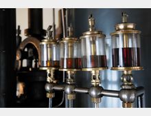 Nahaufnahme von vier Ölerschälern aus Metall mit Glasbehältern, die teilweise mit dunkelrotem Öl gefüllt sind, vor einem metallischen Hintergrund in einer industriellen Maschinenumgebung. Im Hintergrund sind unscharf Maschinenteile zu erkennen.