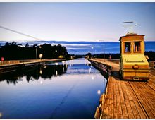 Ein schmaler Kanal mit ruhiger Wasseroberfläche, flankiert von einer hölzernen Plattform rechts und einer parallelen Straße links mit Straßenlaternen. Auf der Plattform steht eine gelbe historische Straßenbahn, und im Hintergrund sind Bäume sowie eine Brücke sichtbar.