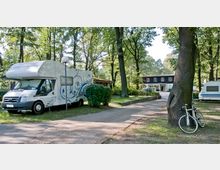 Ein Campingplatz mit einem Wohnmobil und einem geparkten Fahrrad im Vordergrund, umgeben von hohen Bäumen. Im Hintergrund ist ein kleineres Gebäude mit der Aufschrift "Hotel" zu sehen, eingebettet in eine waldreiche Umgebung.