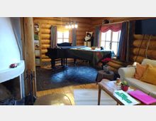 Gemütlicher Innenraum eines Blockhauses mit Holzwänden und Balken. Im Raum befinden sich ein Kamin, ein schwarzer Flügel auf Steinboden, farbig dekorierte Fenster mit Vorhängen, ein heller Sitzbereich mit Kissen sowie ein Couchtisch mit Pflanzen.