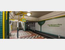 U-Bahn-Station "Franz-Neumann-Platz" mit einer gefliesten Wand gestaltet mit stilisierten Baum-Motiven, grünem Mosaik und Schriftzug des Stationsnamens. Im Vordergrund sind Gleise, ein beleuchtetes gelbes Anzeigesystem und ein Treppenaufgang mit Rolltreppe zu sehen.