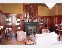 Edles Wohnzimmer mit holzvertäfelten Wänden und einem großen Kamin, der mit detaillierten Schnitzereien verziert ist. Der Raum enthält weiße Polstermöbel, Bücherregale mit Glastüren, eine helle Fliesenbasis und eine große Hängelampe.