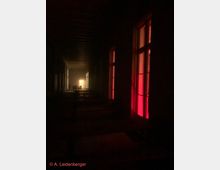 Dunkler Flur mit Holzbänken und schwacher Beleuchtung. Im Hintergrund ist ein heller Lichtschein zu sehen, während die rechten Fenster rot beleuchtet sind.