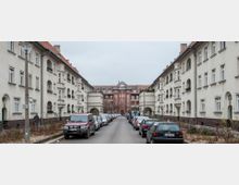 Straßenansicht in einer Wohnsiedlung mit symmetrischen Gebäuden im traditionellen Baustil. Die Häuser haben weiße Fassaden, abgerundete Balkone und rote Dächer; im Hintergrund steht ein großes Backsteingebäude mit Erkern und einem markanten Giebeldach.