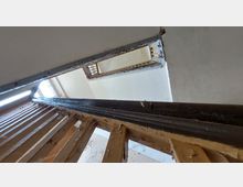 Blick auf ein Treppenhaus in einem alten Gebäude mit abblätternder Farbe an den Holz- und Metallgeländern sowie Sicht auf weiße Wände und ein Fenster, durch das Tageslicht einfällt. Die Perspektive ist nach oben gerichtet und zeigt die Stufen und mehrere Stockwerke.