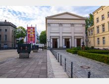 Außenansicht des Maxim-Gorki-Theaters in Berlin, einem neoklassizistischen Gebäude mit Säulenfassade und großen Holztüren. Im Vordergrund ein gepflasterter Weg mit Absperrpfosten, gepflegte Grünanlagen sowie eine Statue und Informationsdisplays; links ein Banner mit Text in mehreren Sprachen.