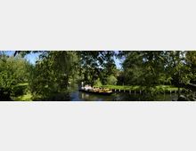 Ein schmaler, bewaldeter Wasserkanal mit einer Holzplattform, auf der Menschen in einem flachen Boot sitzen. Umgeben von dichten Bäumen, Sträuchern und einer Wiese im Hintergrund unter klarem, sonnigem Himmel.